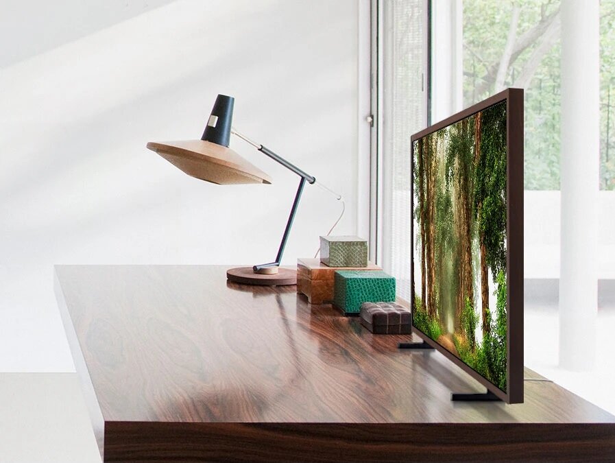 Telewizor Samsung QE32LS03CBUXXH czarny ustawiony na drewnianym biurku stojący obok lampki i pudełek
