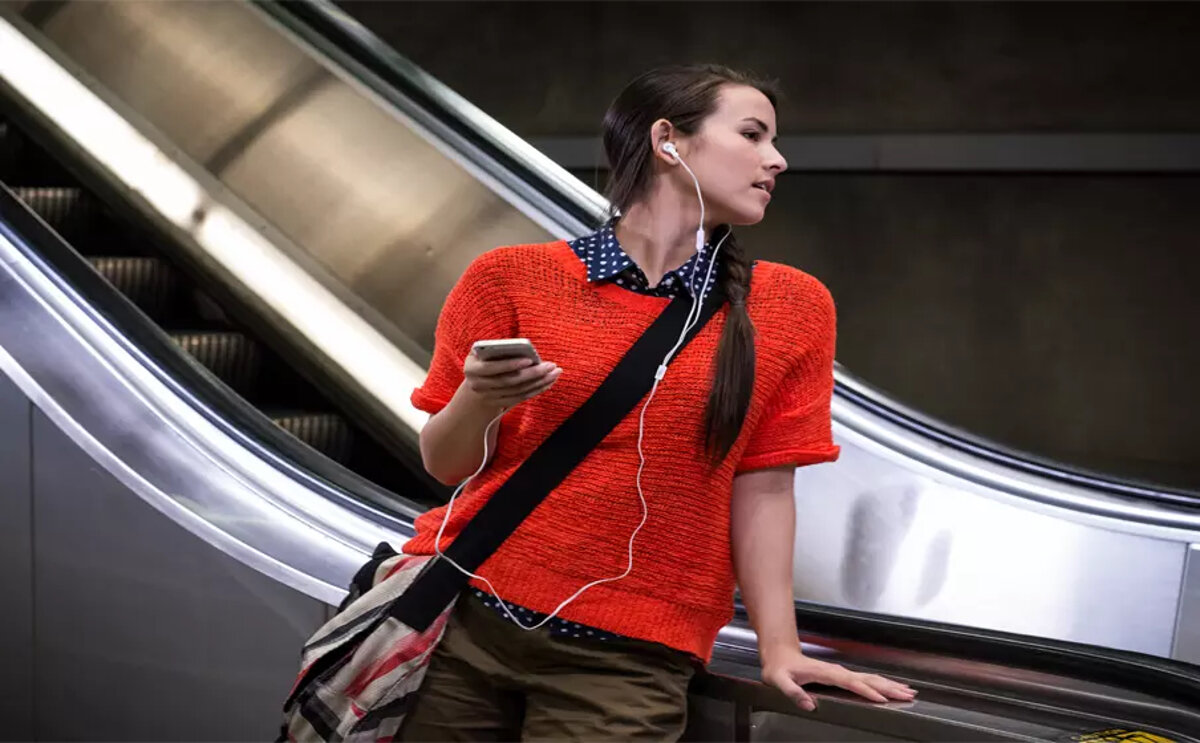 Słuchawki JBL Tune 210 czarne kobieta stojąca na schodach z słuchawkami w uszach w ręku trzyma telefon, który jest podłączony do słuchawek